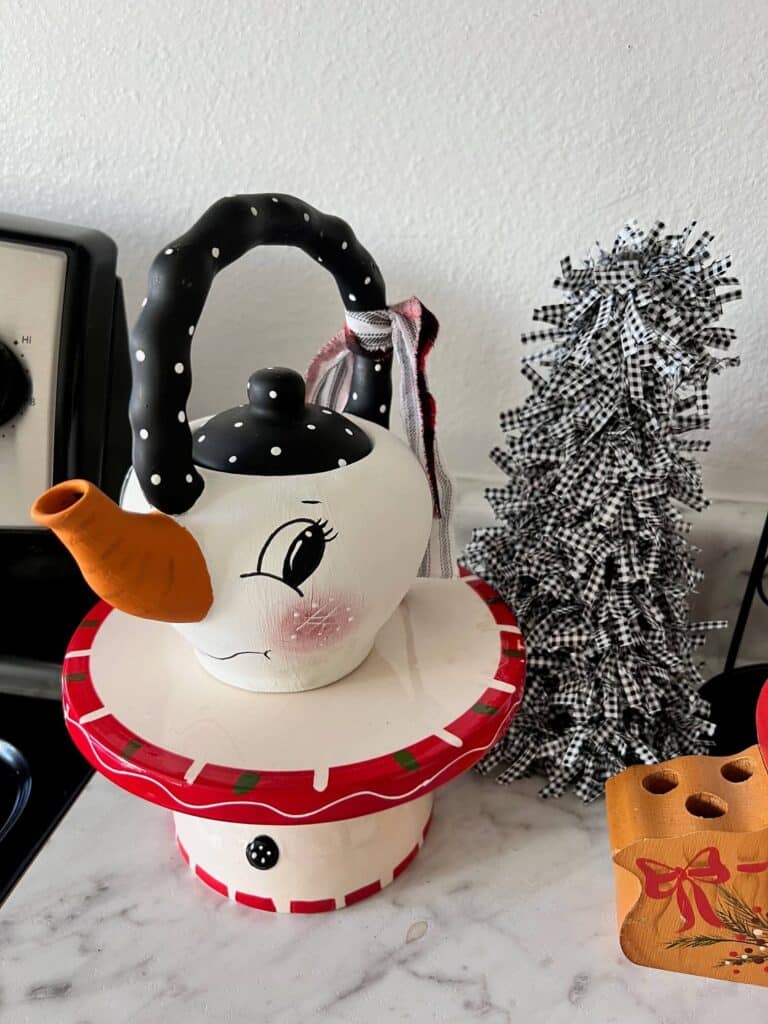 Thrifted Hand painted snowman teapot with the "spout" painted orange as the "carrot" nose, big black eyed, rosy pink cheeks, black and white polka dot handle, and a black, red, and white scrappy fabric bow on the handle.