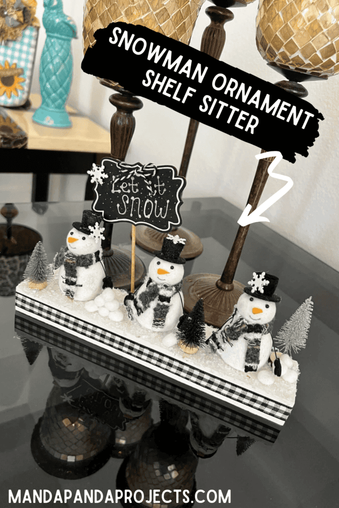 Snowmen Ornament Shelf sitter made with 3 mini felt white snowmen from walmart, on a wood base with a chalkboard sign that says "let it snow", snowballs made with white pom poms, and black and silver bottle brush christmas trees.