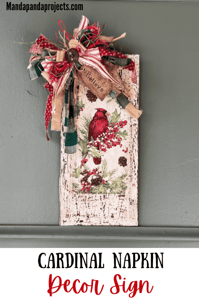 Winter cardinal napkin decoupaged to a distressed white background with a red and green messy bow and a hangtag that says "Believe". Cardinal Napkin Sign