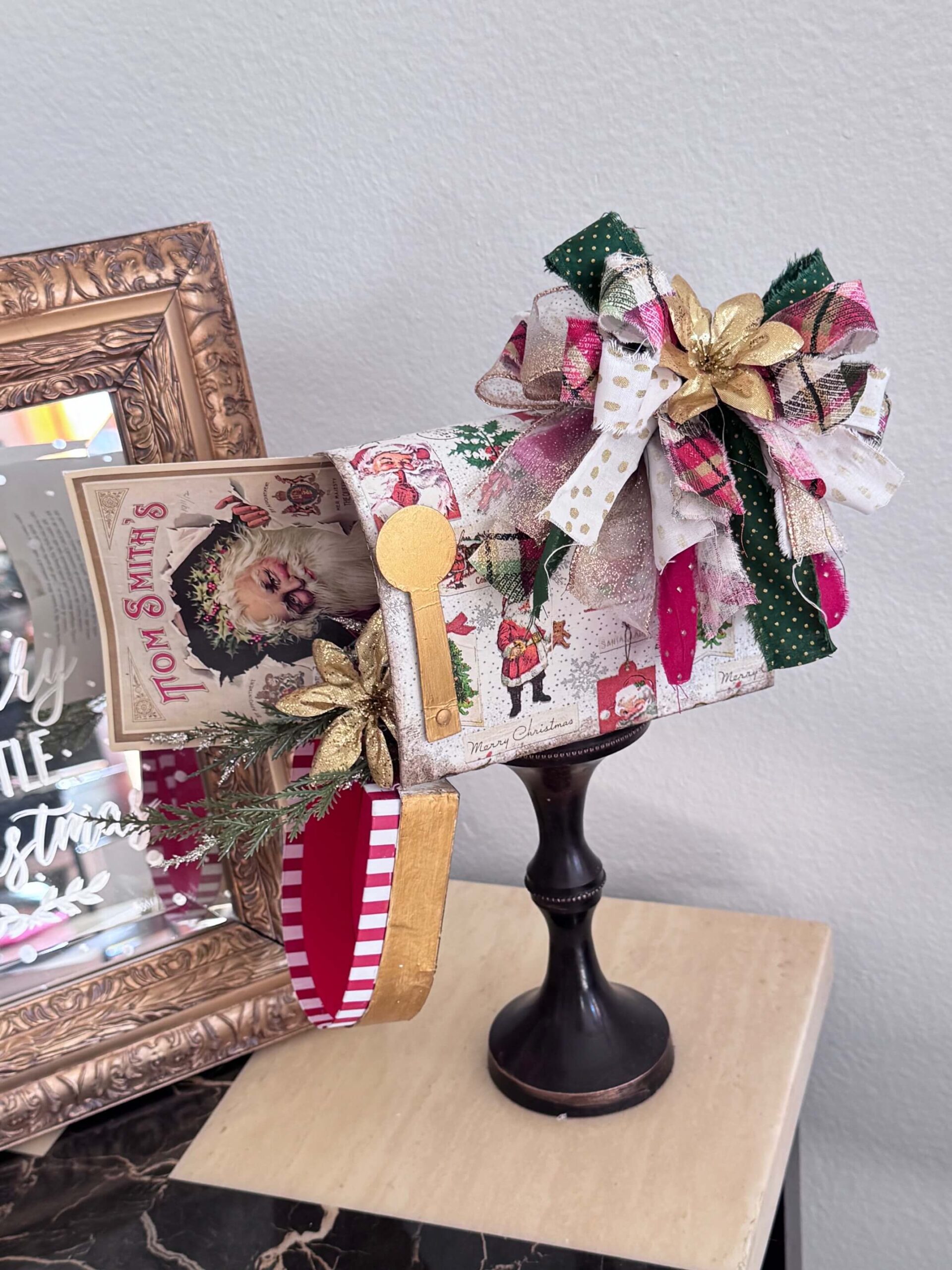 A dollar tree penguin cardboard mailbox that was made over for Christmas with a vintage santa napkin, some gold poinsettias, christmas greenery, and a messy fabric bow on a wood candlestick base for DIY decor. 