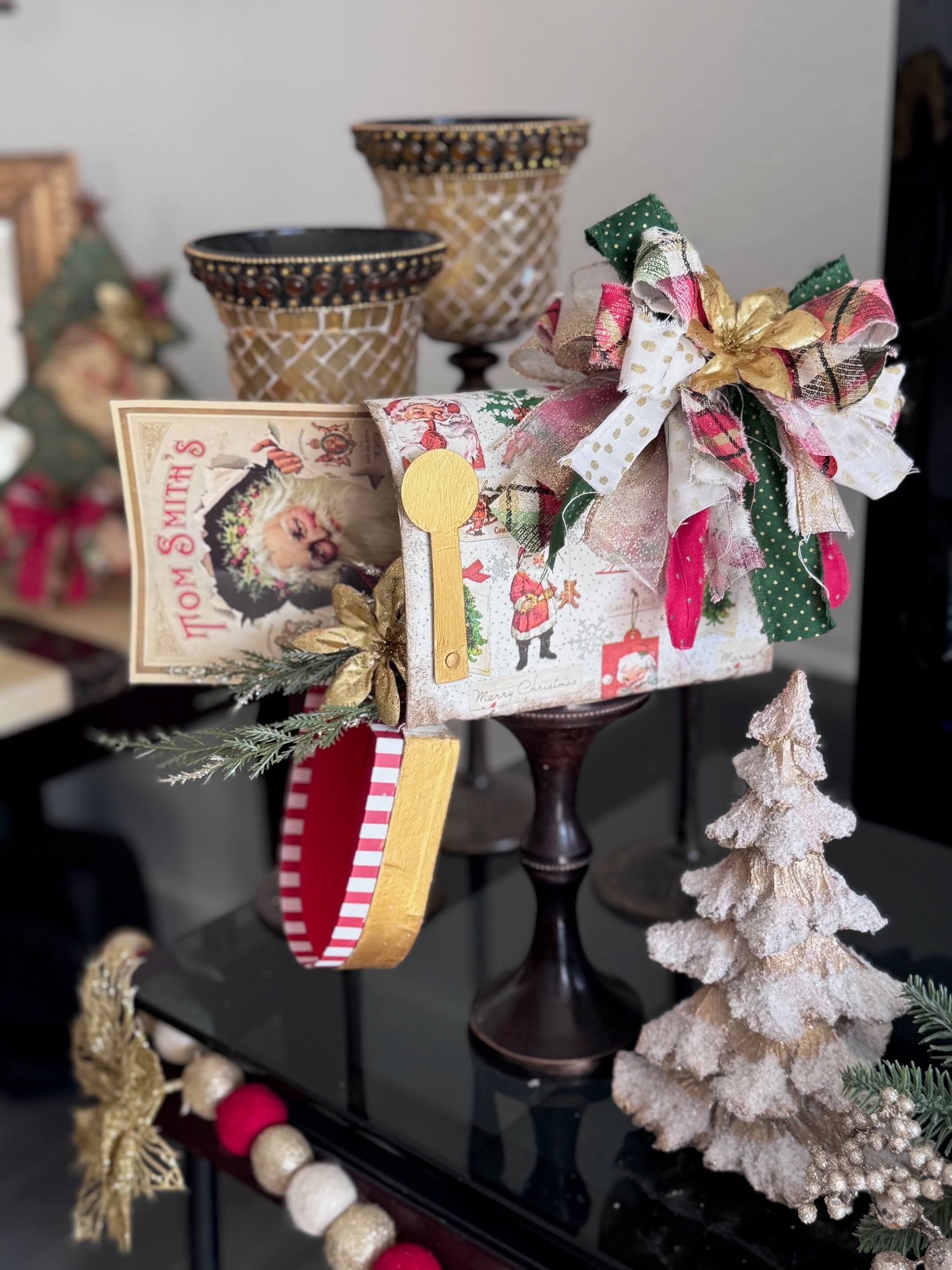 A dollar tree penguin cardboard mailbox that was made over for Christmas with a vintage santa napkin, some gold poinsettias, christmas greenery, and a messy fabric bow on a wood candlestick base for DIY decor. 