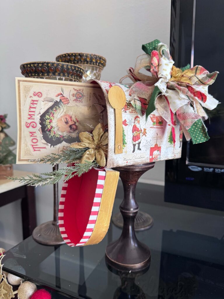 A dollar tree penguin cardboard mailbox that was made over for Christmas with a vintage santa napkin, some gold poinsettias, christmas greenery, and a messy fabric bow on a wood candlestick base for DIY decor. 