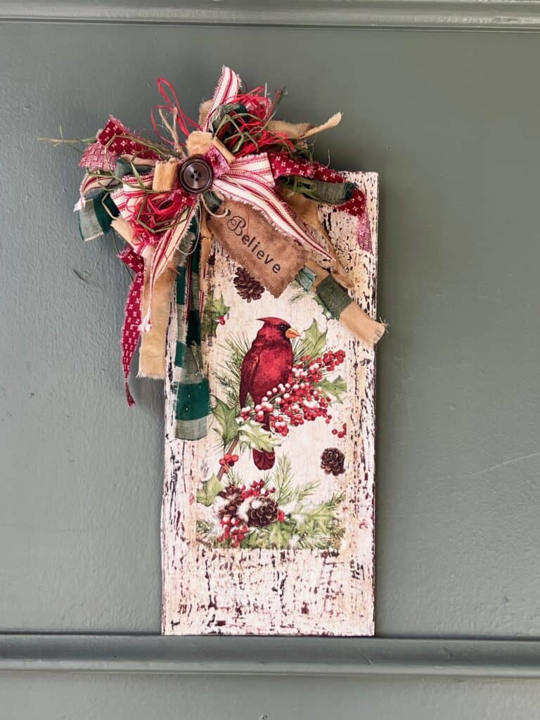 Winter cardinal napkin decoupaged to a distressed white background with a red and green messy bow and a hangtag that says "Believe". Cardinal Napkin Sign