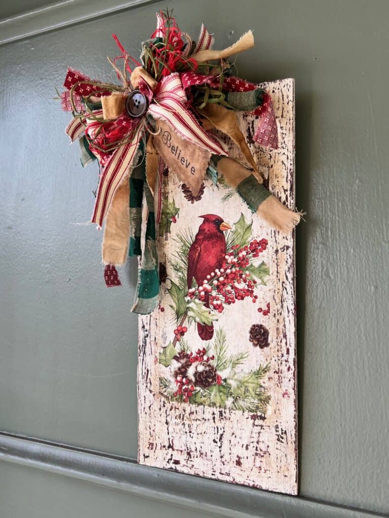 Winter cardinal napkin decoupaged to a distressed white background with a red and green messy bow and a hangtag that says "Believe". Cardinal Napkin Sign