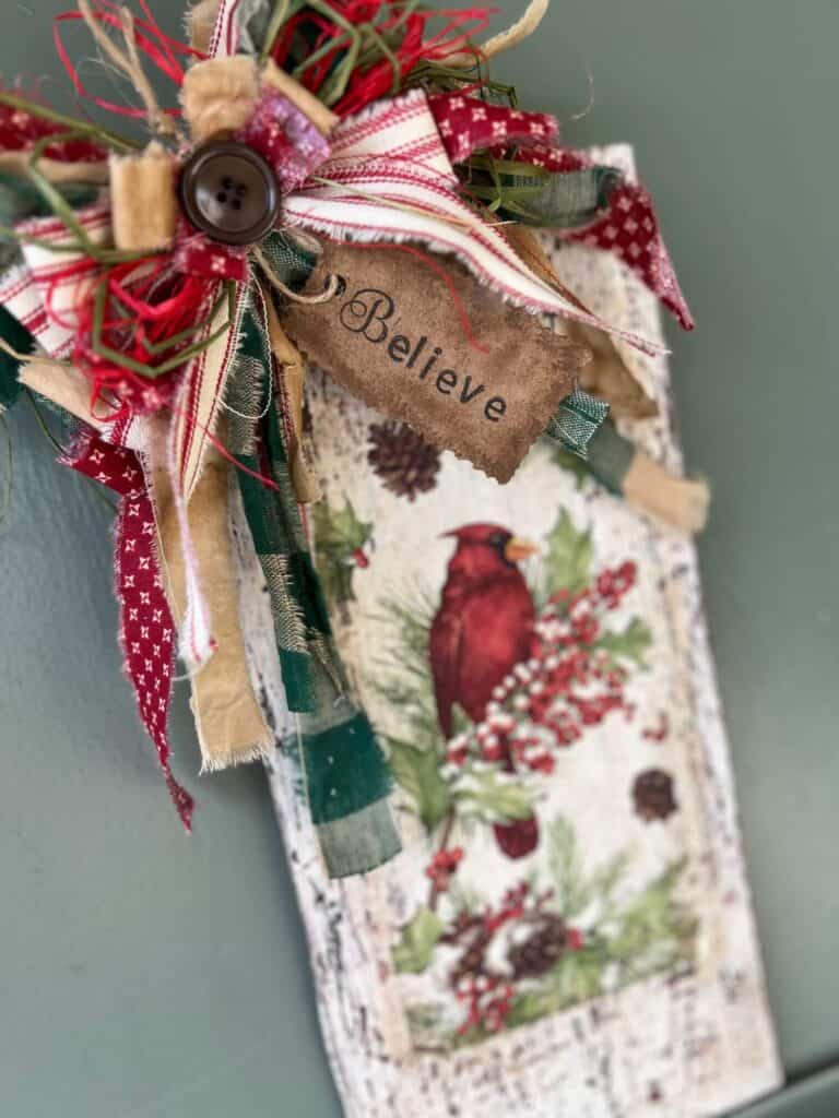 Winter cardinal napkin decoupaged to a distressed white background with a red and green messy bow and a hangtag that says "Believe". Cardinal Napkin Sign