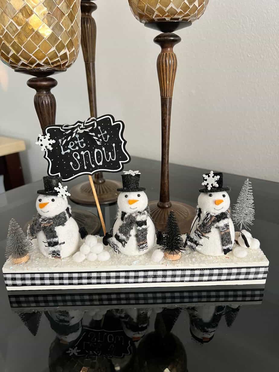 Snowmen Ornament Shelf sitter made with 3 mini felt white snowmen from walmart, on a wood base with a chalkboard sign that says "let it snow", snowballs made with white pom poms, and black and silver bottle brush christmas trees.