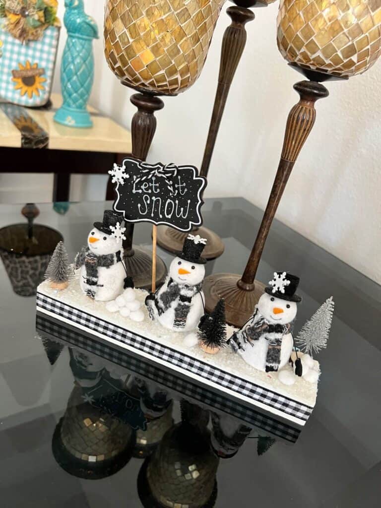 Snowmen Ornament Shelf sitter made with 3 mini felt white snowmen from walmart, on a wood base with a chalkboard sign that says "let it snow", snowballs made with white pom poms, and black and silver bottle brush christmas trees.