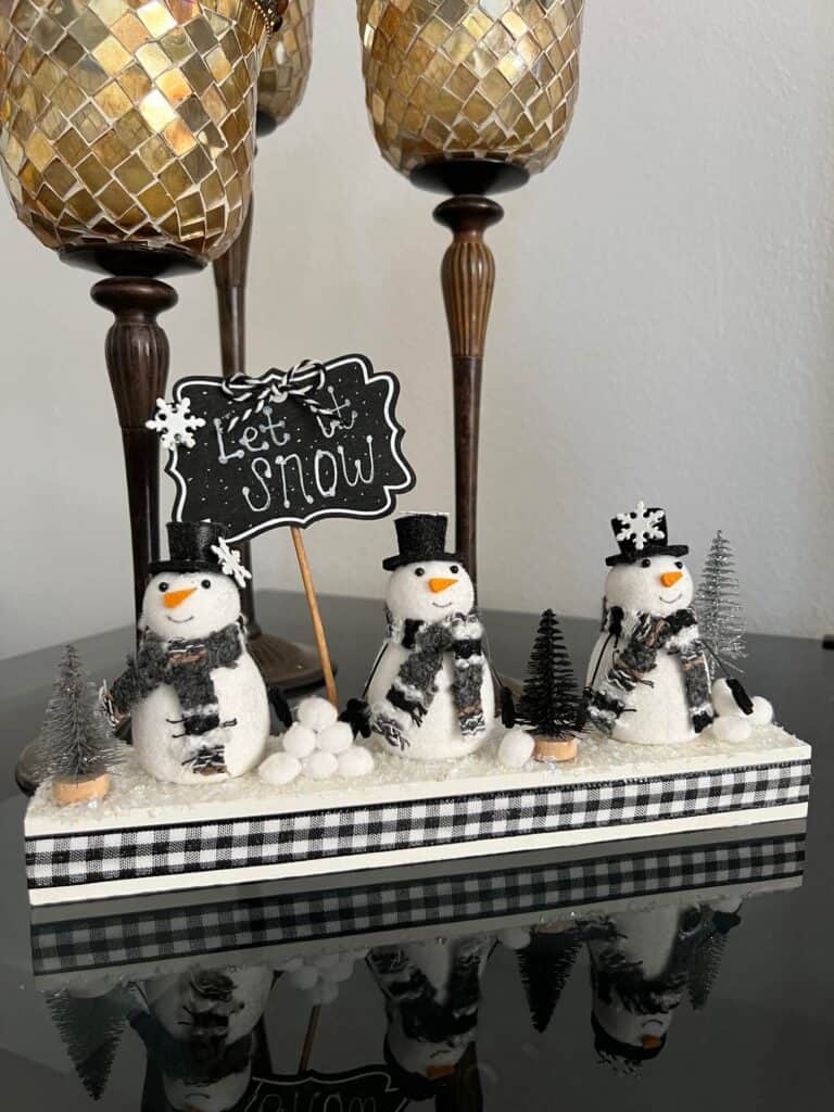 Snowmen Ornament Shelf sitter made with 3 mini felt white snowmen from walmart, on a wood base with a chalkboard sign that says "let it snow", snowballs made with white pom poms, and black and silver bottle brush christmas trees.