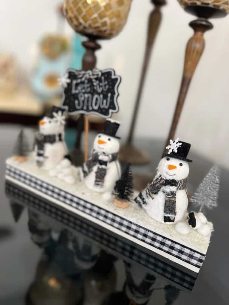 Snowmen Ornament Shelf sitter made with 3 mini felt white snowmen from walmart, on a wood base with a chalkboard sign that says "let it snow", snowballs made with white pom poms, and black and silver bottle brush christmas trees.