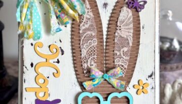 Corrugated cardboard bunny with messy fabric bow with purple, teal and yellow, with teal heart shaped glasses, a heart nose, butterflies, flowers, and a Dollar Tree bunny Hop sign, on a chippy white background.