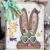 Corrugated cardboard bunny with messy fabric bow with purple, teal and yellow, with teal heart shaped glasses, a heart nose, butterflies, flowers, and a Dollar Tree bunny Hop sign, on a chippy white background.