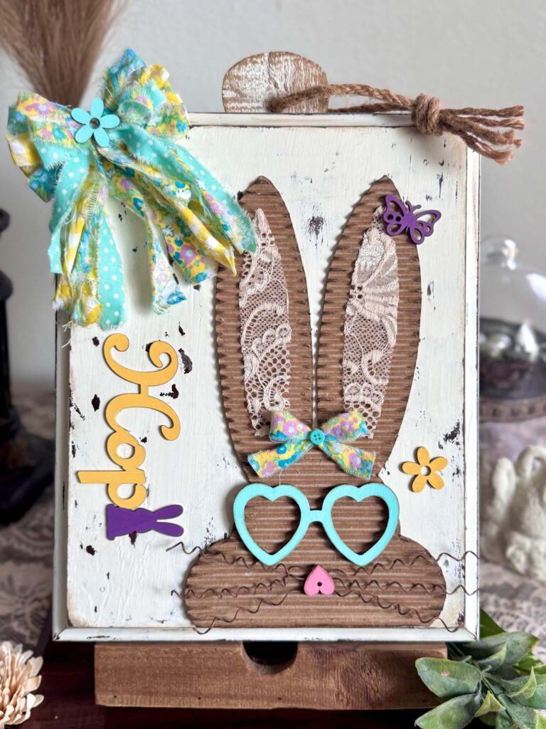 Corrugated cardboard bunny with messy fabric bow with purple, teal and yellow, with teal heart shaped glasses, a heart nose, butterflies, flowers, and a Dollar Tree bunny Hop sign, on a chippy white background.