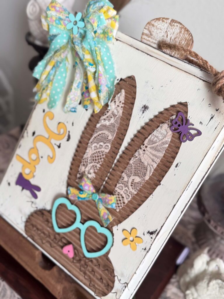 Corrugated cardboard bunny with messy fabric bow with purple, teal and yellow, with teal heart shaped glasses, a heart nose, butterflies, flowers, and a Dollar Tree bunny Hop sign, on a chippy white background.