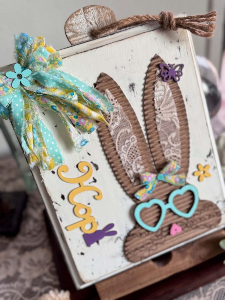 Corrugated cardboard bunny with messy fabric bow with purple, teal and yellow, with teal heart shaped glasses, a heart nose, butterflies, flowers, and a Dollar Tree bunny Hop sign, on a chippy white background.