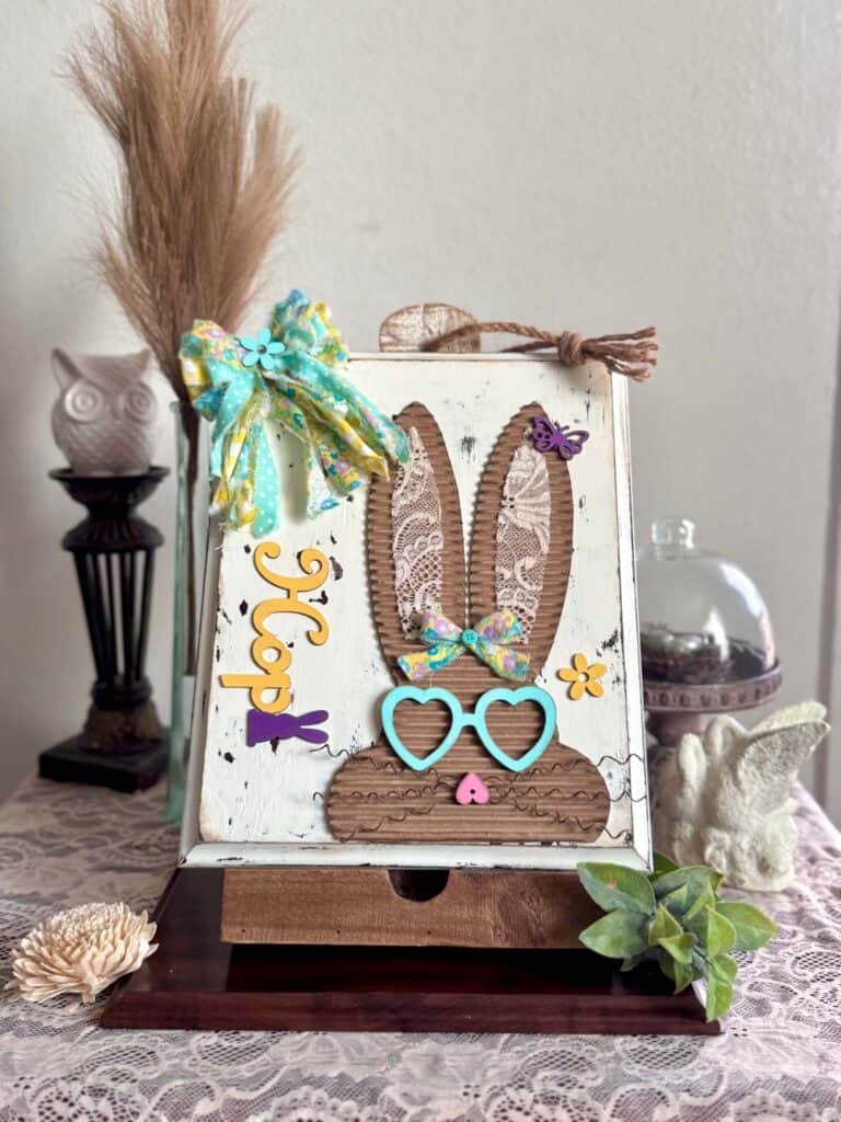 Corrugated cardboard bunny with messy fabric bow with purple, teal and yellow, with teal heart shaped glasses, a heart nose, butterflies, flowers, and a Dollar Tree bunny Hop sign, on a chippy white background.