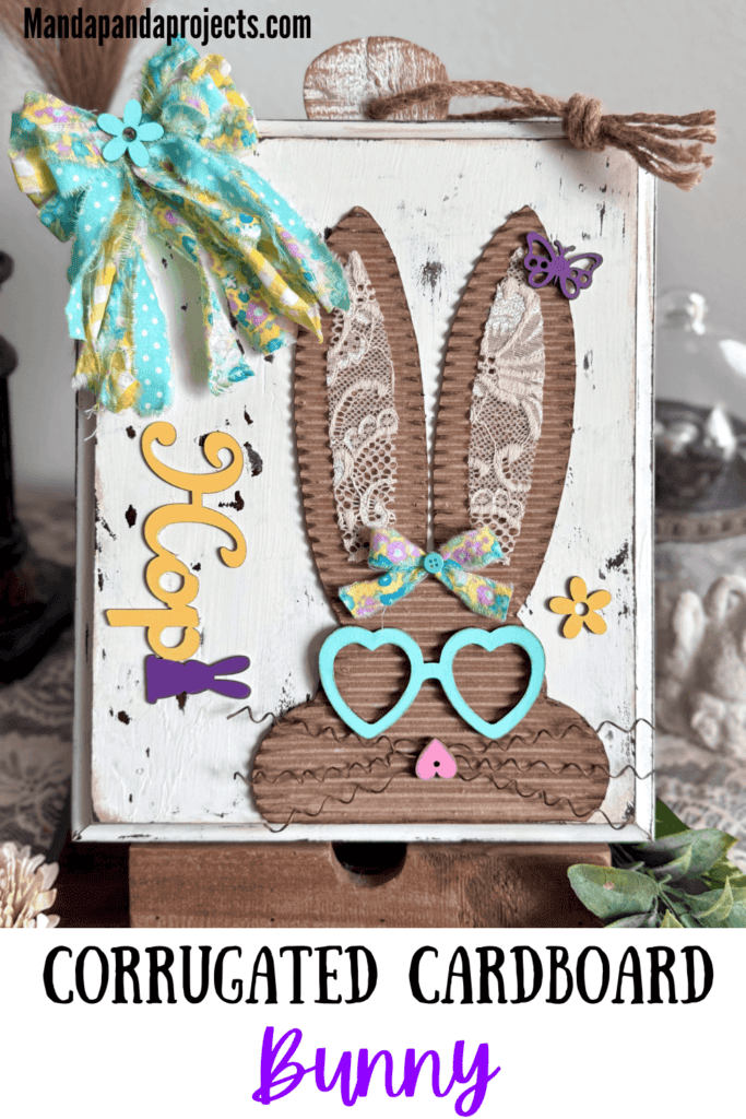 Corrugated cardboard bunny with messy fabric bow with purple, teal and yellow, with teal heart shaped glasses, a heart nose, butterflies, flowers, and a Dollar Tree bunny Hop sign, on a chippy white background.