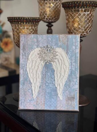 A Dollar Tree stretch canvas with pretty distressed blue scrapbook paper, a textured set of angel wings made with joint compound, sparkling with diamond dust and a totally dazzled bling in the center of the wings. A loving memory craft.