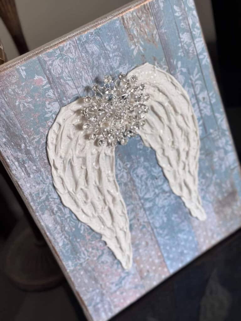 A Dollar Tree stretch canvas with pretty distressed blue scrapbook paper, a textured set of angel wings made with joint compound, sparkling with diamond dust and a totally dazzled bling in the center of the wings. A loving memory craft.