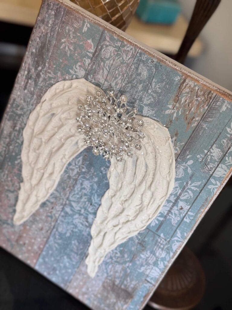 A Dollar Tree stretch canvas with pretty distressed blue scrapbook paper, a textured set of angel wings made with joint compound, sparkling with diamond dust and a totally dazzled bling in the center of the wings. A loving memory craft.