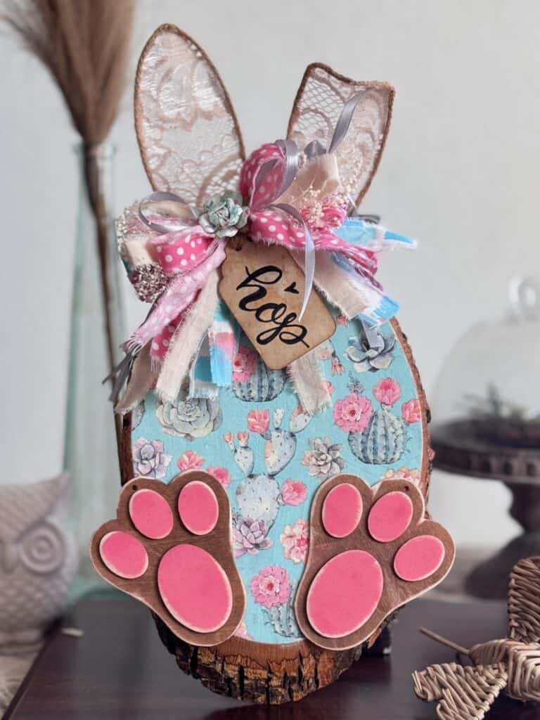 Thrifted wood round decoupaged with a succulent floral spring napkin to look like a bunny with Jute twine ears, a messy fabric bow, a hangtag that says "Hop" and pink bunny feet for diy Spring and easter crafts and decor. 