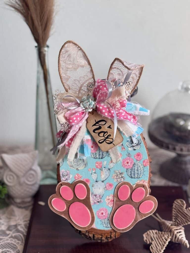 Thrifted wood round decoupaged with a succulent floral spring napkin to look like a bunny with Jute twine ears, a messy fabric bow, a hangtag that says "Hop" and pink bunny feet for diy Spring and easter crafts and decor. 