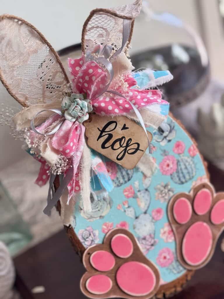 Thrifted wood round decoupaged with a succulent floral spring napkin to look like a bunny with Jute twine ears, a messy fabric bow, a hangtag that says "Hop" and pink bunny feet for diy Spring and easter crafts and decor. 