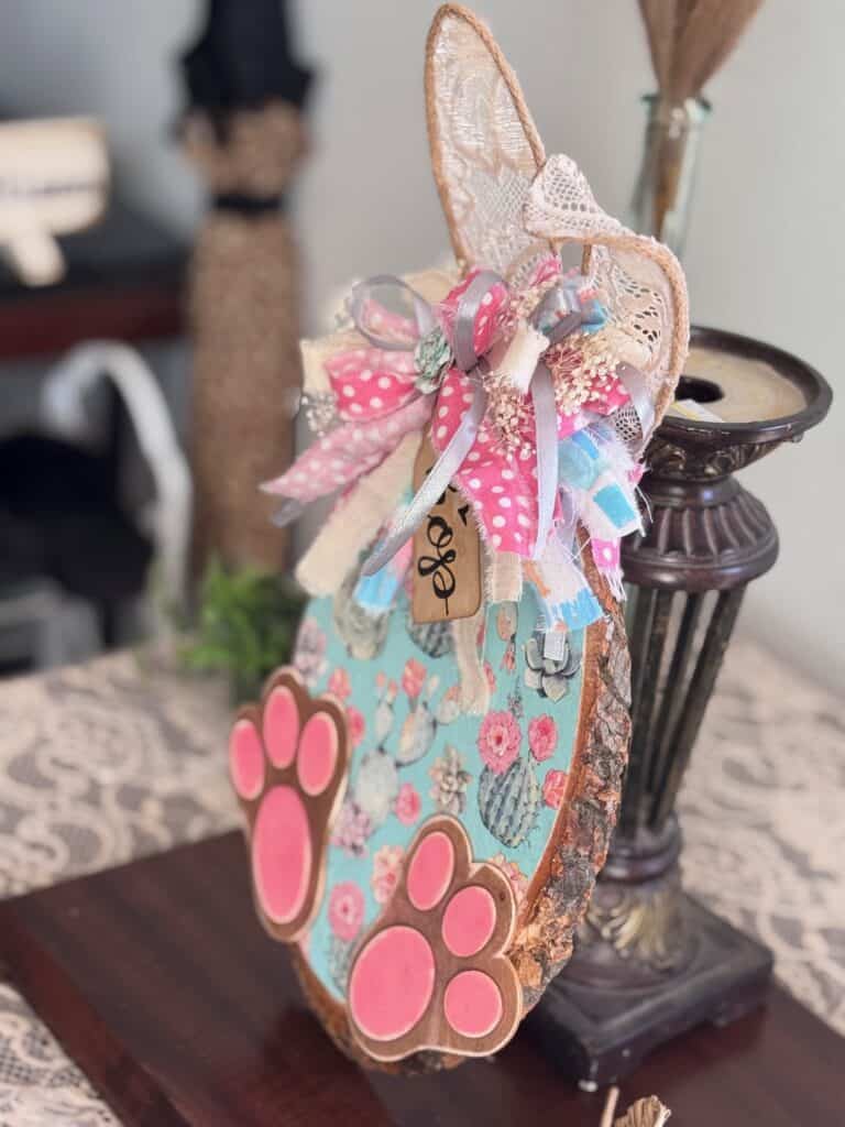 Thrifted wood round decoupaged with a succulent floral spring napkin to look like a bunny with Jute twine ears, a messy fabric bow, a hangtag that says "Hop" and pink bunny feet for diy Spring and easter crafts and decor. 