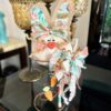An Easter and Spring bunny made with a rusty bedspring, a muslin head, burlap ears, and a floral colorful bow with wood carrot embellishments and dried white flowers.