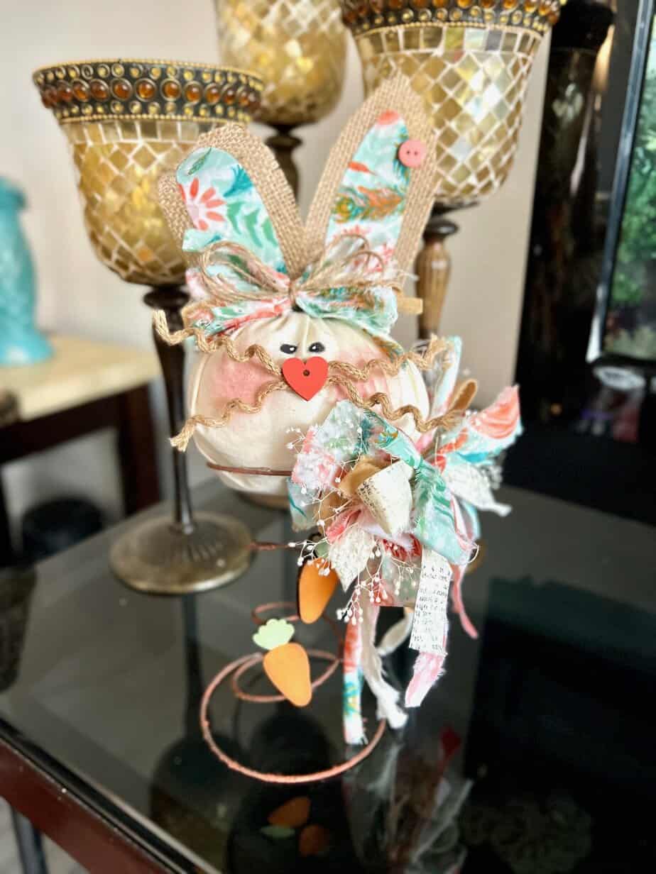 An Easter and Spring bunny made with a rusty bedspring, a muslin head, burlap ears, and a floral colorful bow with wood carrot embellishments and dried white flowers.