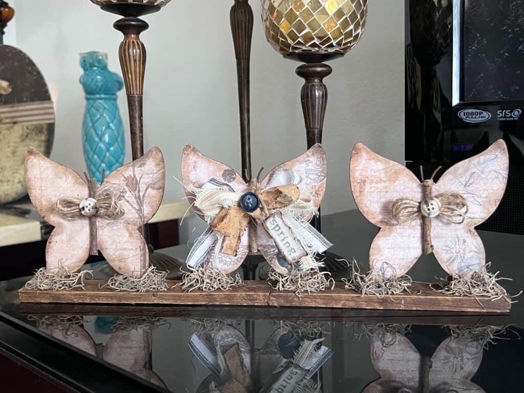 3 Wood Butterfly shelf sitter with a vintage vibe with a spring hangtag and twine and fabric bows, sitting on a base with spanish moss.