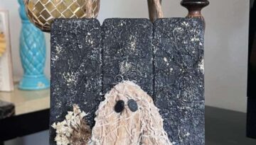 Coffee stained Muslin creepy cloth ghost holding a creme natural dried flower bouquet with a twine bow and rusty bell, with a black background with a gold starry night and a jute rope hanger.