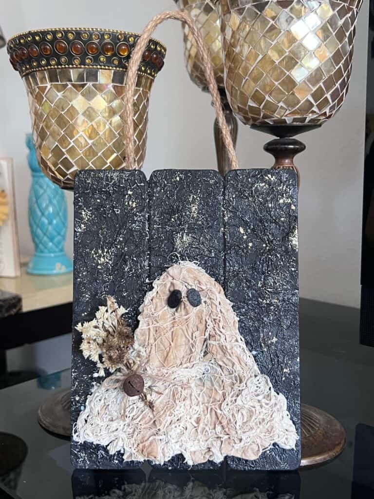 Coffee stained Muslin creepy cloth ghost holding a creme natural dried flower bouquet with a twine bow and rusty bell, with a black background with a gold starry night and a jute rope hanger.