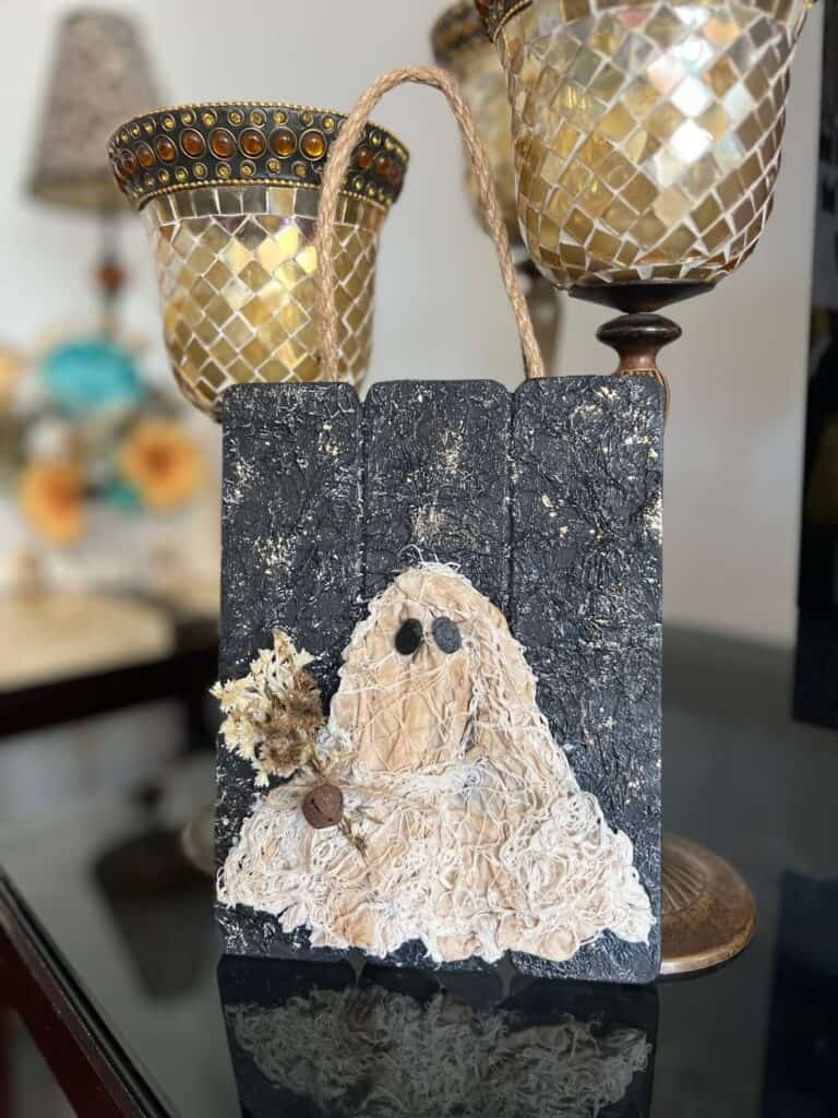 Coffee stained Muslin creepy cloth ghost holding a creme natural dried flower bouquet with a twine bow and rusty bell, with a black background with a gold starry night and a jute rope hanger.