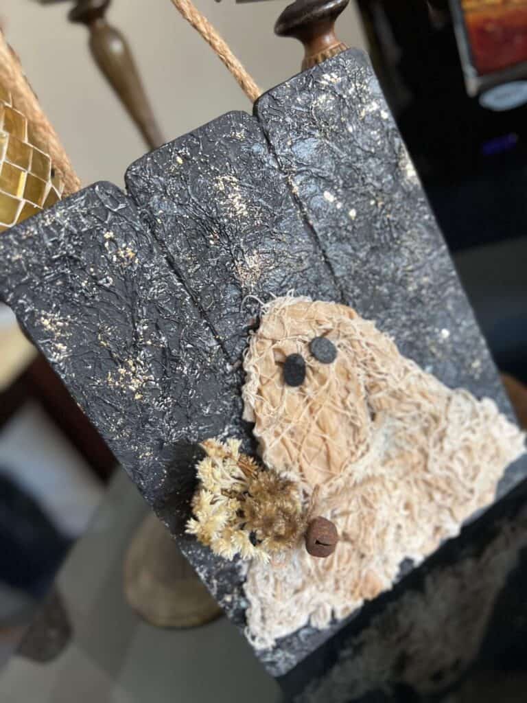 Coffee stained Muslin creepy cloth ghost holding a creme natural dried flower bouquet with a twine bow and rusty bell, with a black background with a gold starry night and a jute rope hanger.