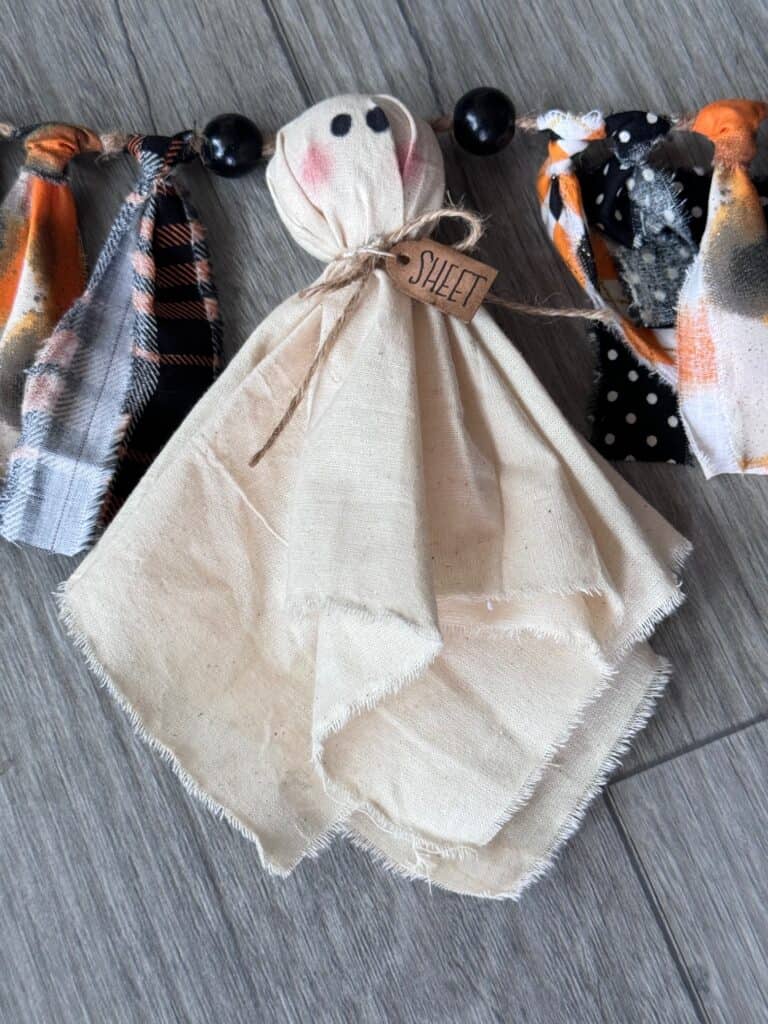 Muslin ghost with a hangtag around its neck that says "SHEET" and orange and black fabric on each side.
