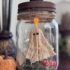 Floating Ghost in a mason Jar made from muslin and creepy cloth and hanging by fishing line with a tea light at the top to give it a spooky cute halloween vibe. The bottom has green reindeer moss, a gravestone that says RIP and a small pumpkin.