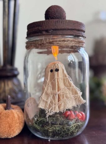 Floating Ghost in a mason Jar made from muslin and creepy cloth and hanging by fishing line with a tea light at the top to give it a spooky cute halloween vibe. The bottom has green reindeer moss, a gravestone that says RIP and a small pumpkin.