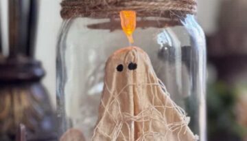 Floating Ghost in a mason Jar made from muslin and creepy cloth and hanging by fishing line with a tea light at the top to give it a spooky cute halloween vibe. The bottom has green reindeer moss, a gravestone that says RIP and a small pumpkin.