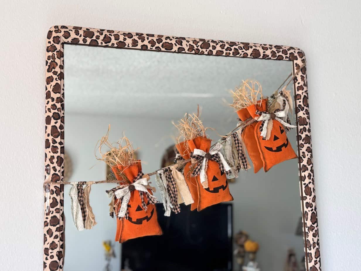 Dollar Tree Pumpkin Treat Bag Garland with 3 pumpkins hanging on twine and raffia sticking out the top, strung across a mirror with a leopard print border for dall decor.