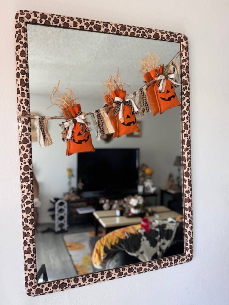 Dollar Tree Pumpkin Treat Bag Garland with 3 pumpkins hanging on twine and raffia sticking out the top, strung across a mirror with a leopard print border for dall decor.