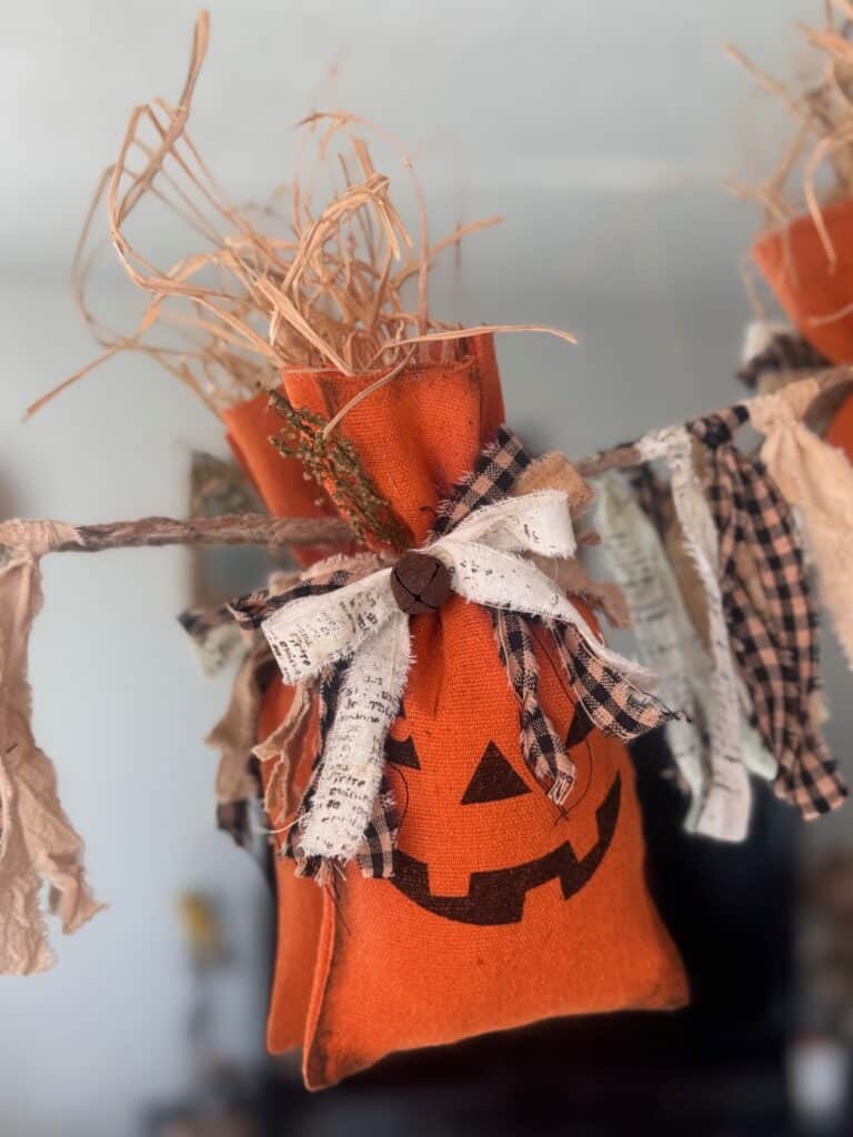 Dollar Tree Pumpkin Treat Bag Garland with raffia sticking out the top, a homespun bow with a rusty bell and sweet annie in the bow. 