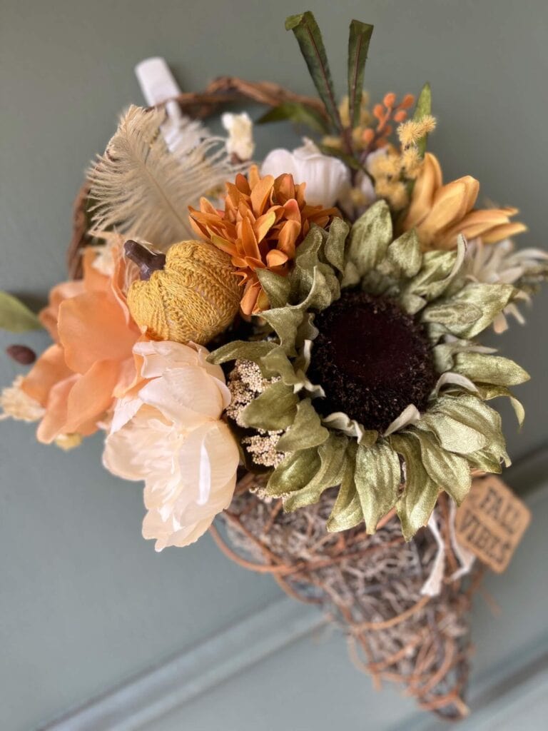 Fall floral grapevine wreath basket door decor hanging on a sage green front door with pretty fall flowers, a sweater pumpkin pick and a hangtag that says "Fall vibes".