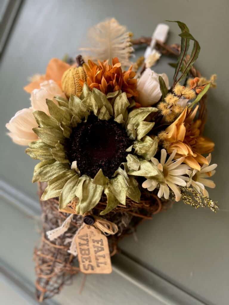 Fall floral grapevine wreath basket door decor hanging on a sage green front door with pretty fall flowers, a sweater pumpkin pick and a hangtag that says "Fall vibes".