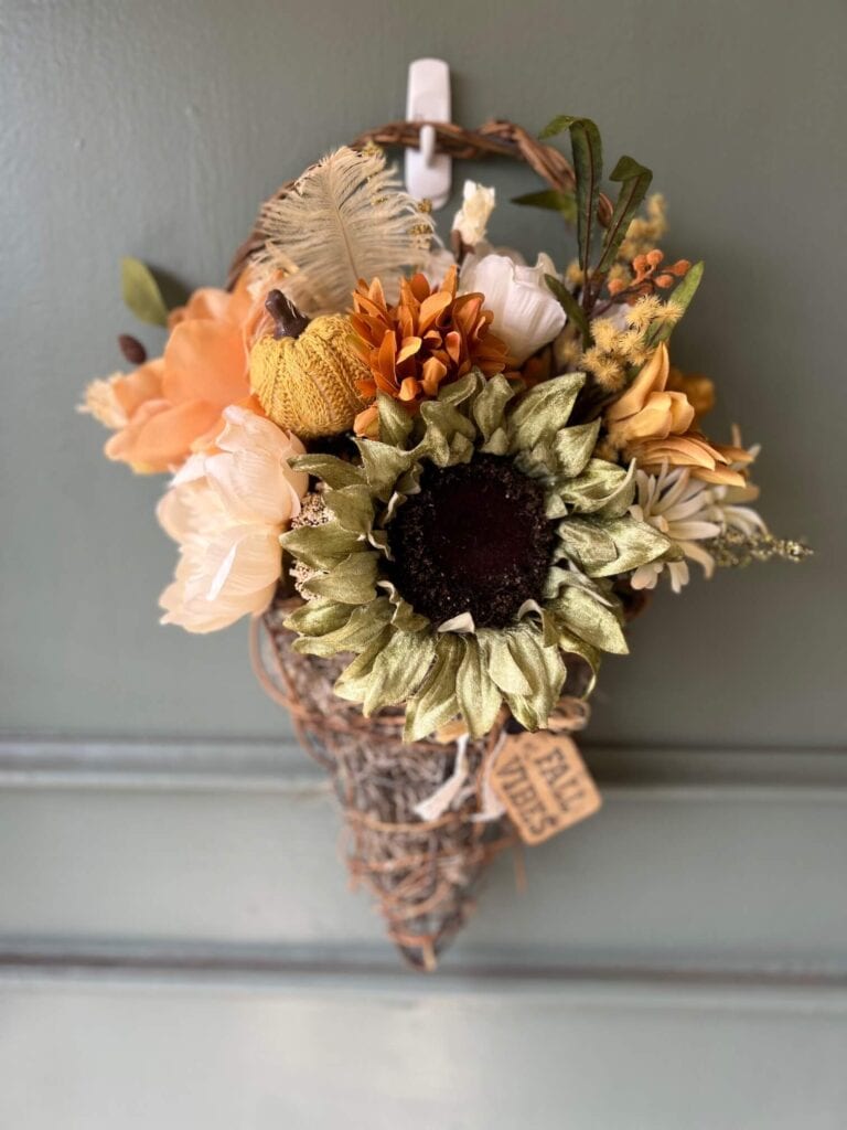 Fall floral grapevine wreath basket door decor hanging on a sage green front door with pretty fall flowers, a sweater pumpkin pick and a hangtag that says "Fall vibes".