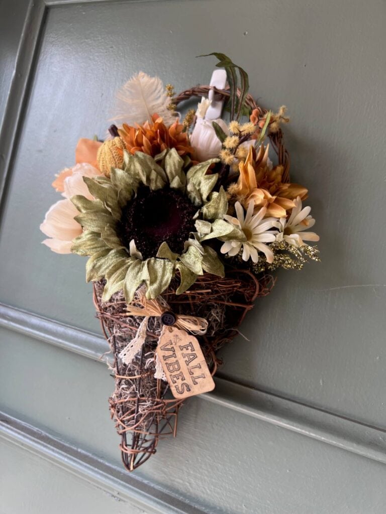 Fall floral grapevine wreath basket door decor hanging on a sage green front door with pretty fall flowers, a sweater pumpkin pick and a hangtag that says "Fall vibes".